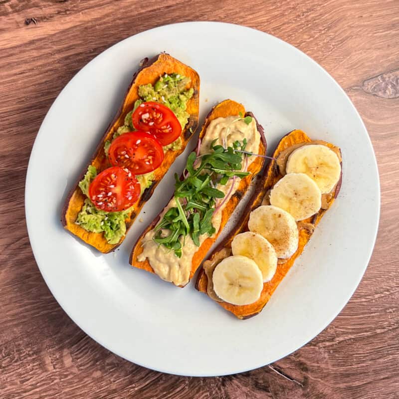 Three sweet potato toasts on a plate, each with different toppings: avocado and tomato; hummus and arugula; peanut butter and banana.