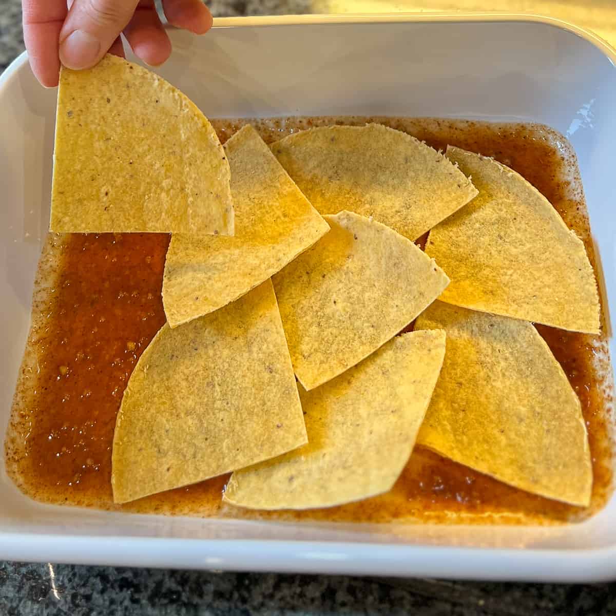 top view of corn tortilla slices being arranged in casserole dish on top of red sauce