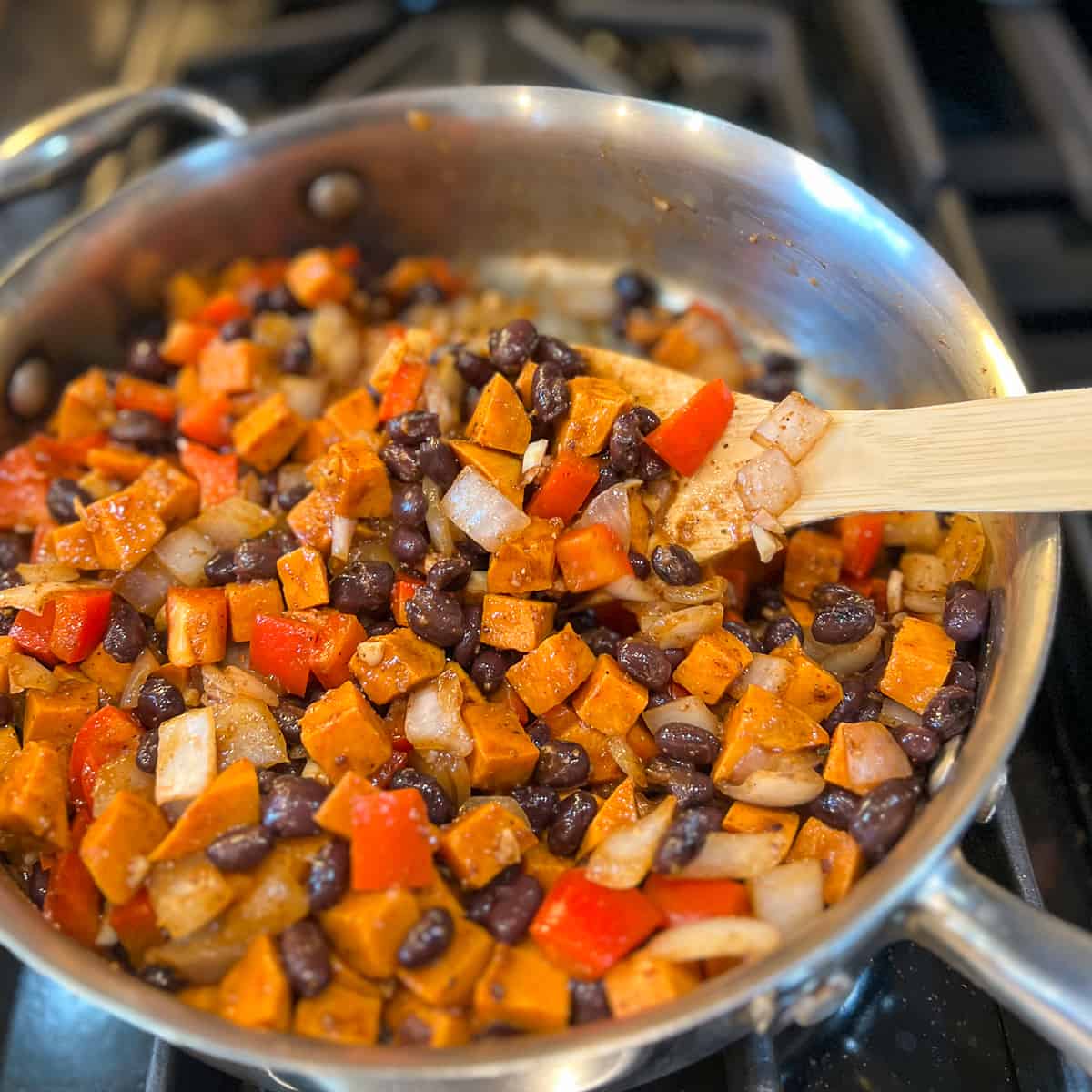 a wooden spoon stirring the veggies, beans and sweet potato with sauce