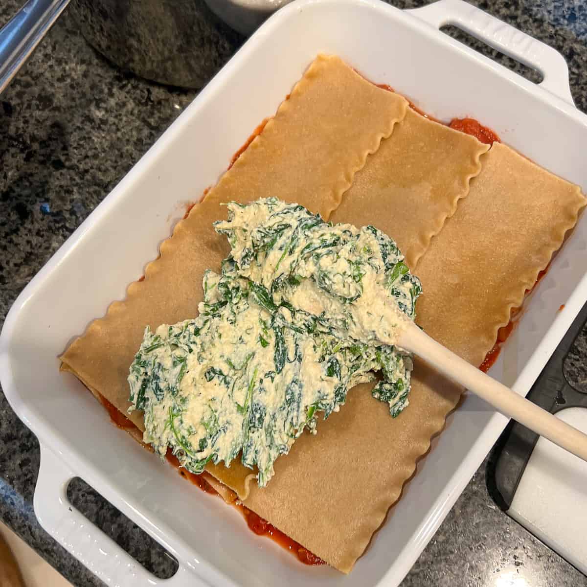 lasagna being assembled with a layer of pasta and ricotta spinach filling being spread across the pasta layer in a white rectangular baking dish