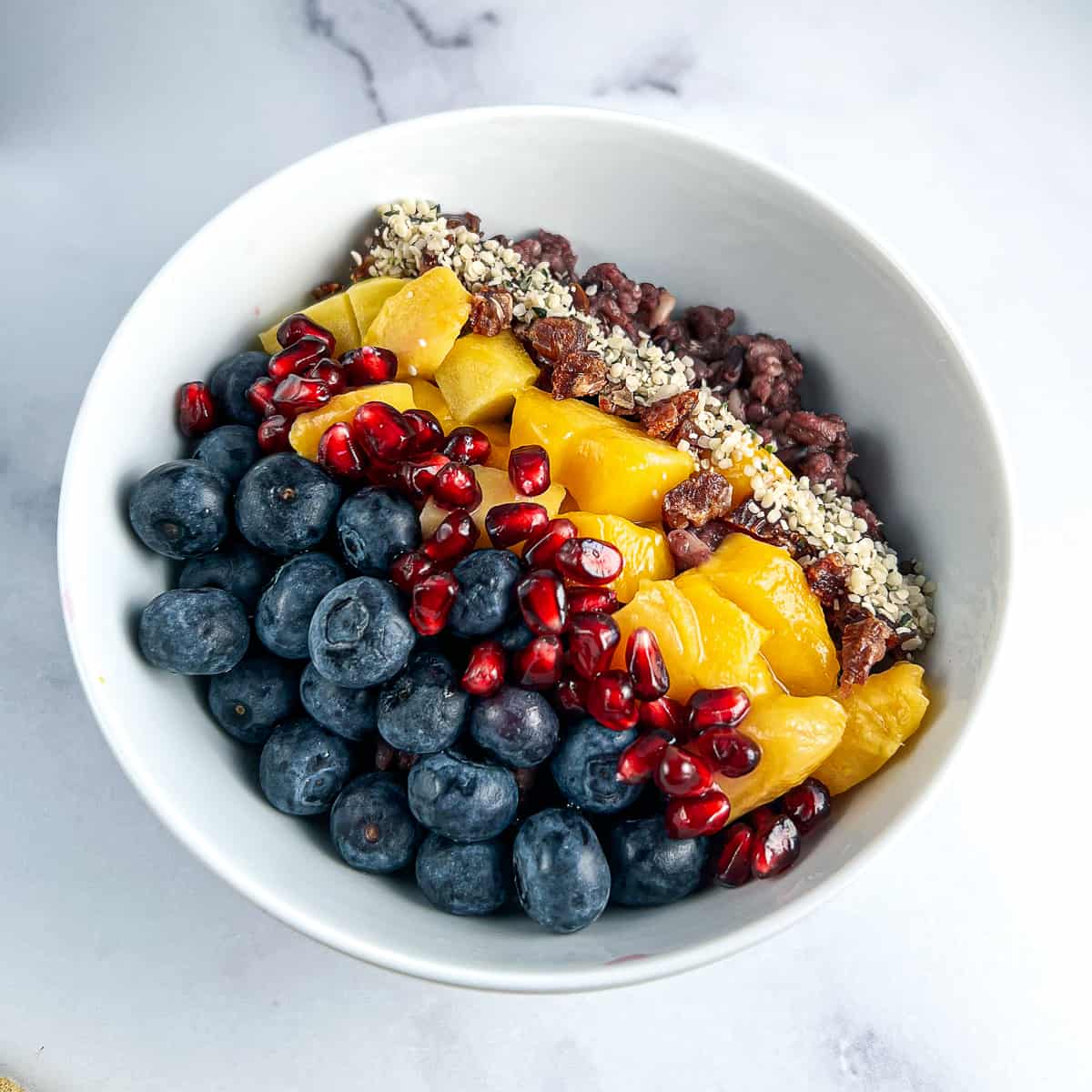 top view close up of ancient grains bowl with fresh blueberries, pomegranate seeds, mango, dates, hemp hearts in a white bowl