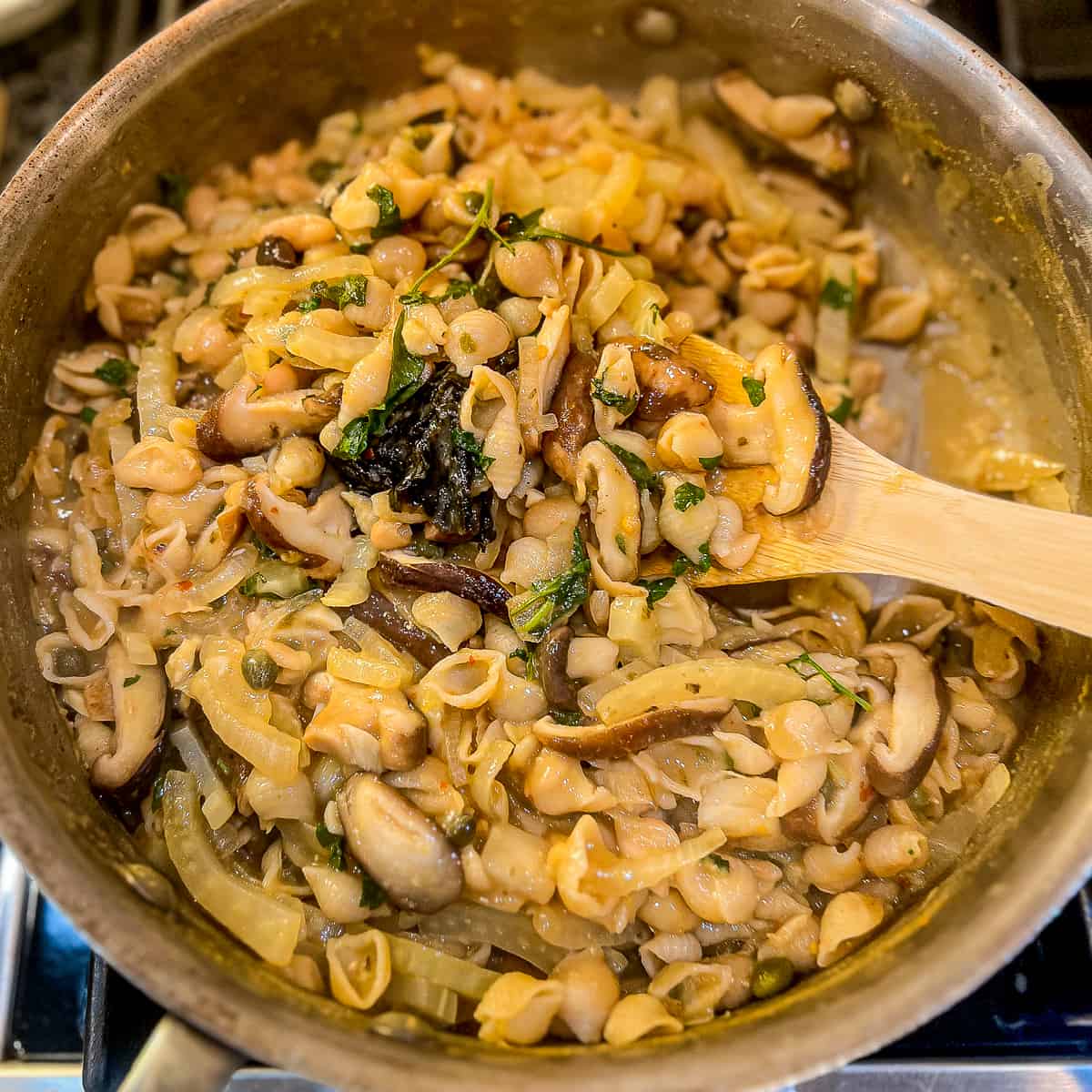 top view close up of a sauté pan with vegan mushroom pasta on the stovetop and a wooden spoon dishing up a scoop