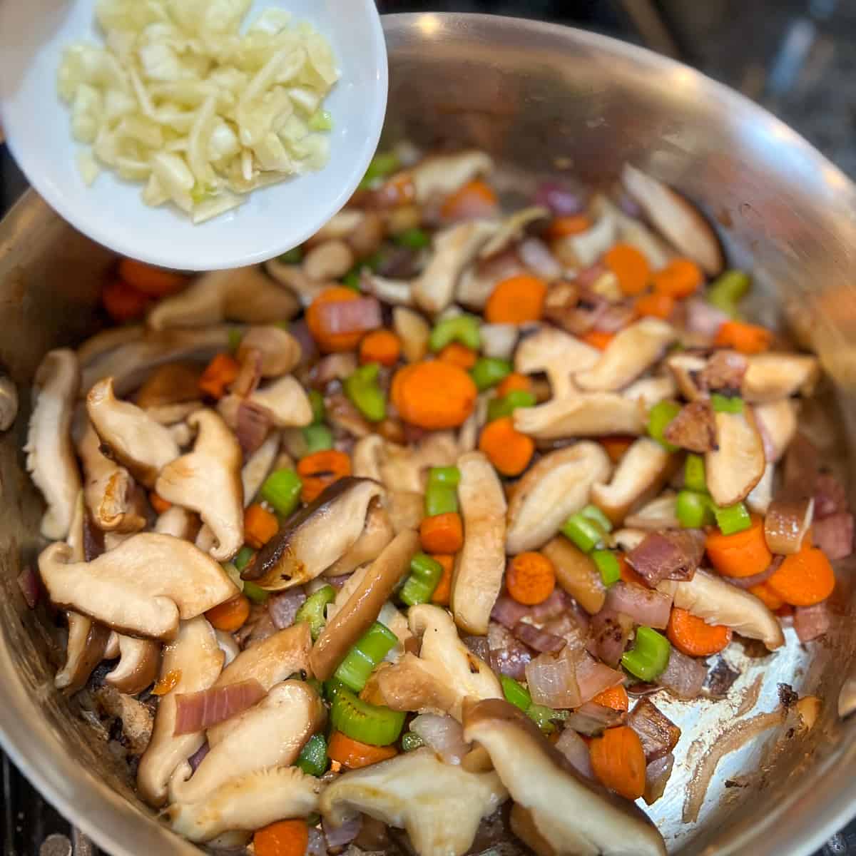 top view of minced garlic being added to a large sauté pan with cooked mushrooms and veggies.