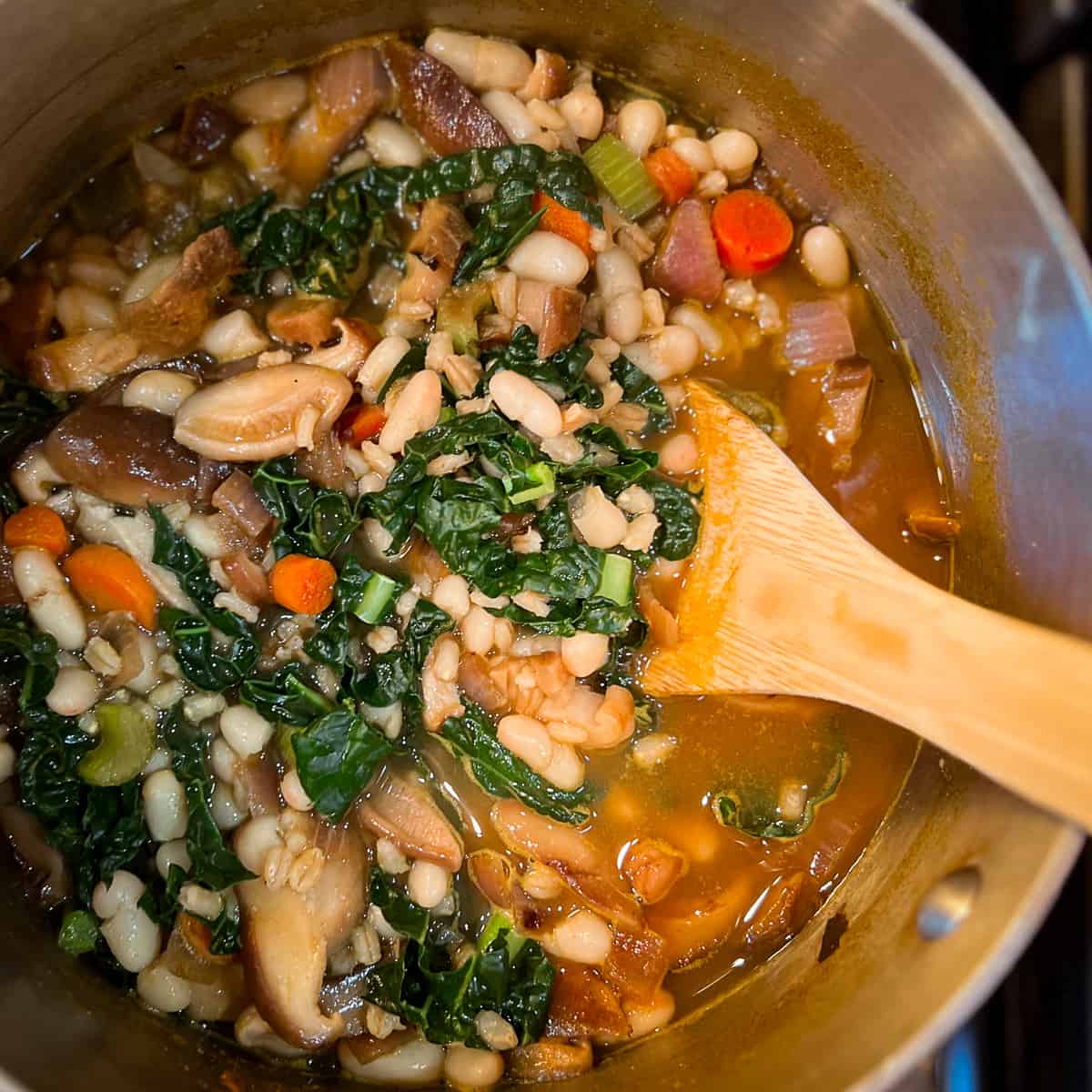 top view close up of a pot of mushroom barley soup with kale and cannellini beans and wooden spoon sticking out of the pot