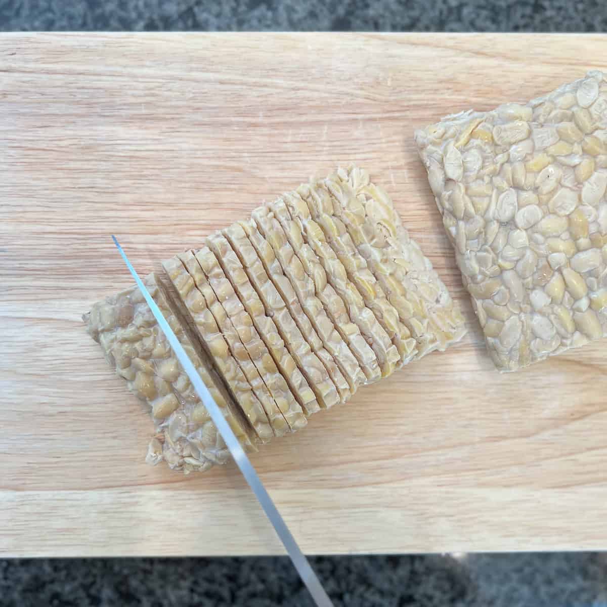 a tempeh block being sliced into bacon strips with a knife on a wooden cutting board