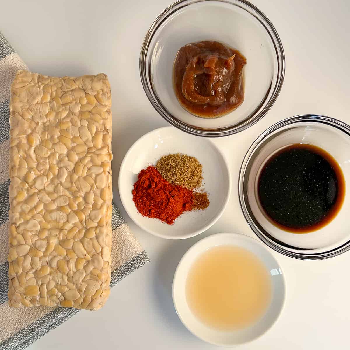 top view of the key ingredients for tempeh bacon: block of tempeh, simple homemade date syrup, coconut aminos, apple cider vinegar, and spices