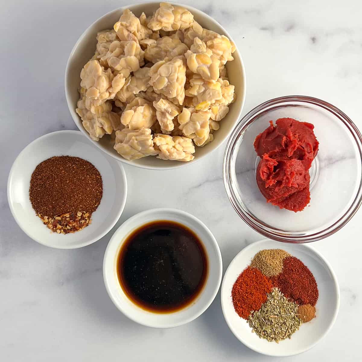 Top view ingredients for tempeh chorizo: tempeh, tomato paste, smoked and sweet paprika, oregano, cinnamon, cumin, chili powder, crushed red pepper, coconut aminos