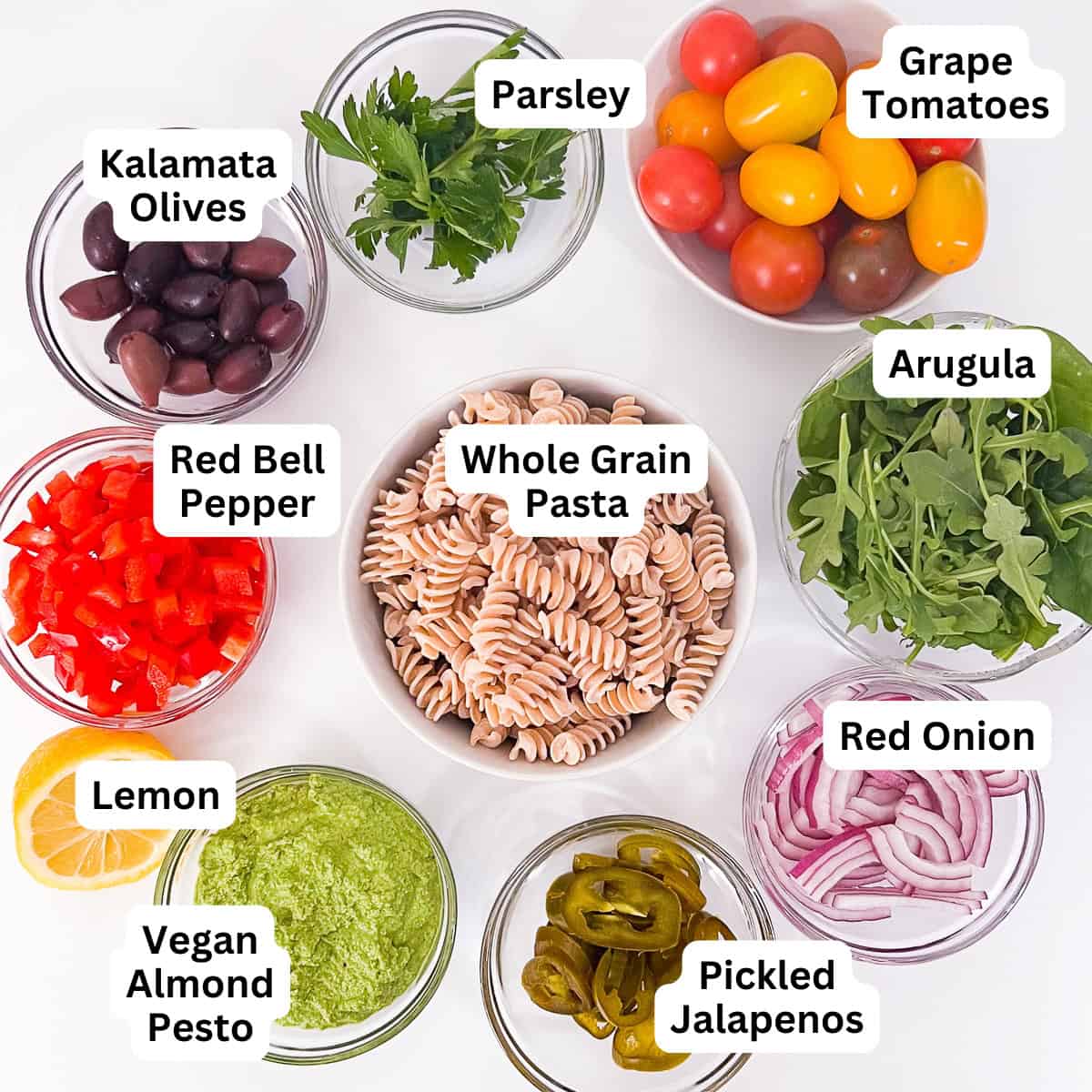 Ingredients for vegan pesto pasta salad: whole grain pasta, olives, parsley, grape tomatoes, arugula, red bell pepper, red onion, pickled jalapenos, Lemon, vegan almond pesto.