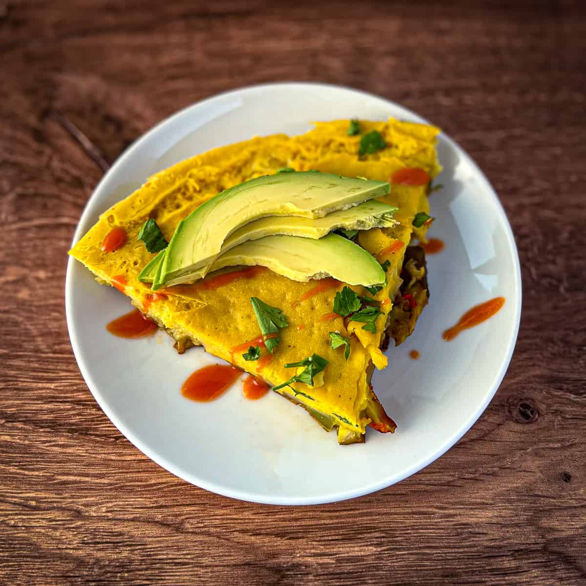 top view close up of a slice of vegan frittata topped with sliced avocado and hot sauce sitting on a white plate