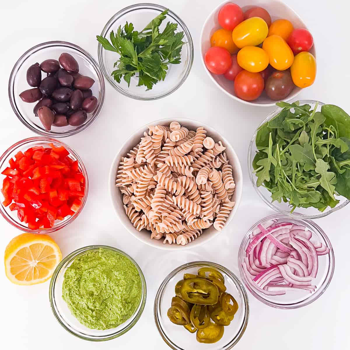 top view of the key ingredients: whole grain fusilli pasta, vegan almond pesto, cherry/grape tomatoes, arugula/spinach, red onion, pickled jalapenos, red bell pepper, pitted Kalamata olives, lemon, fresh parsley
