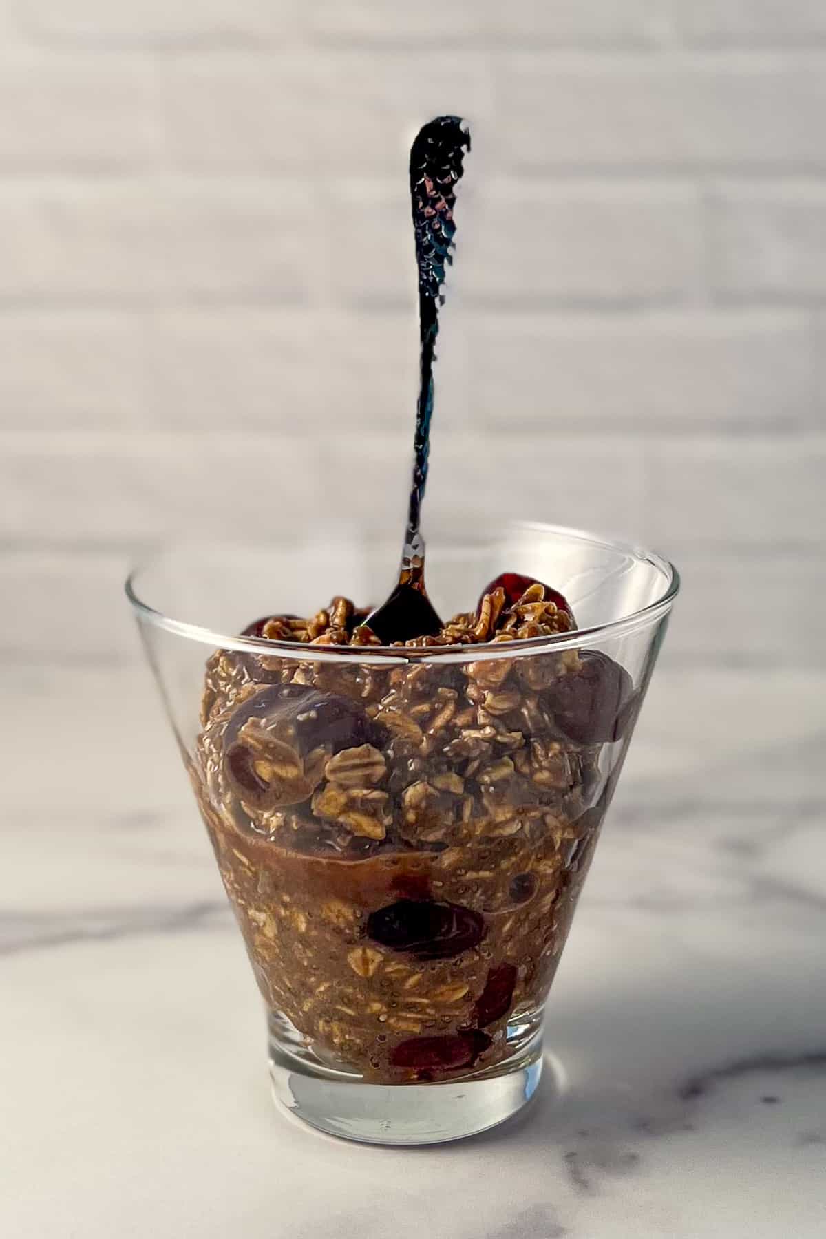 side view close up of black forest overnight oats in a glass dish with a spoon sticking out 