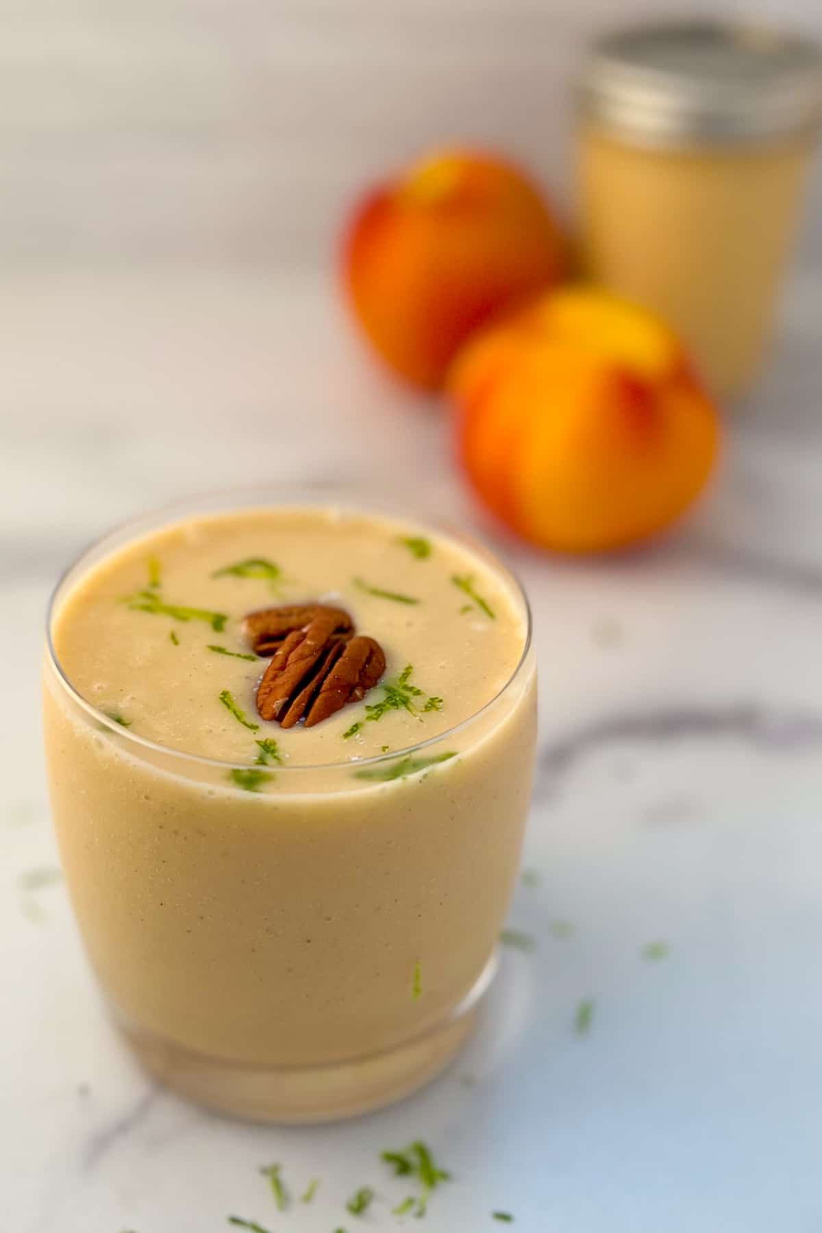 side view of a glass with peach banana smoothie with lime zest and pecans; peaches and mason jar blurred in the background