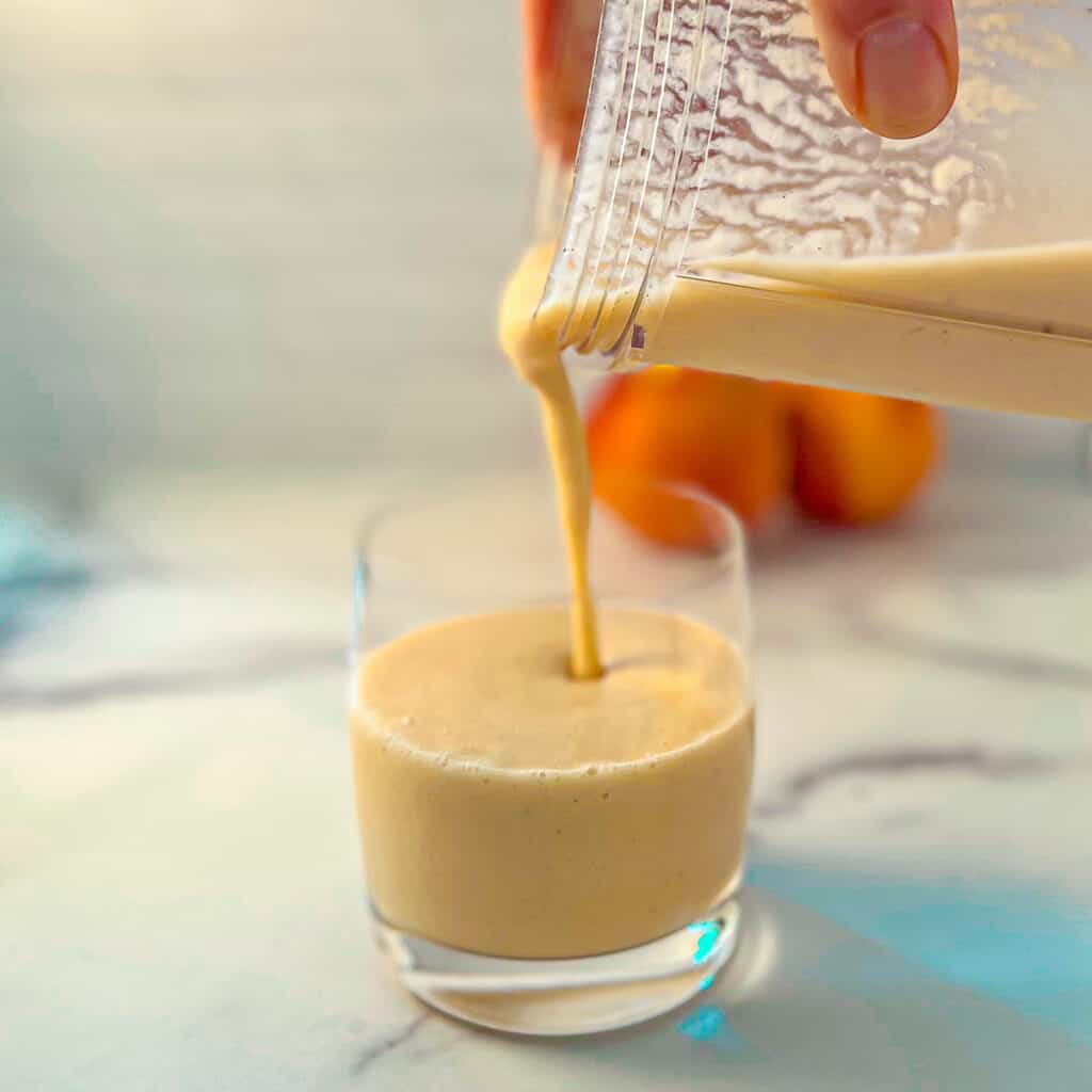 side view close up of peach banana smoothie being poured into a glass from a blender; two peaches blurred in the background