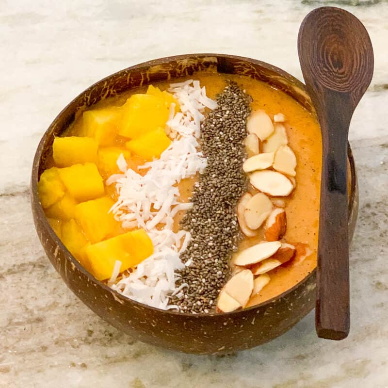 top view of tropical smoothie bowl in a wooden bowl with wooden spoon; chopped pineapple, shredded coconut, chia seeds and sliced almonds as toppings