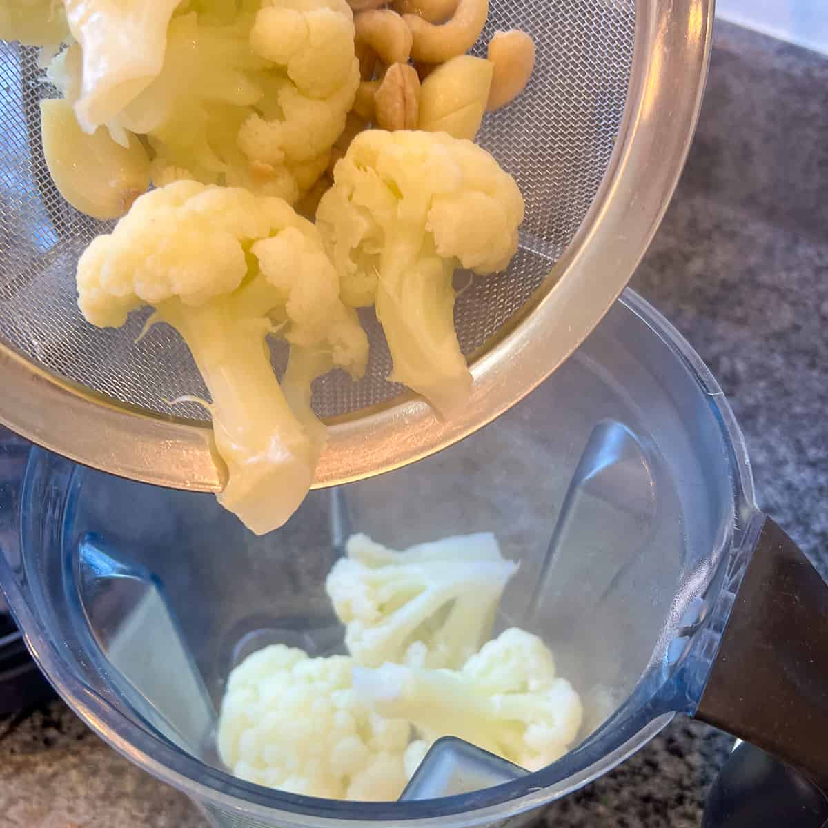 top view of cooked cauliflower, cashews and garlic cloves being added to a blender