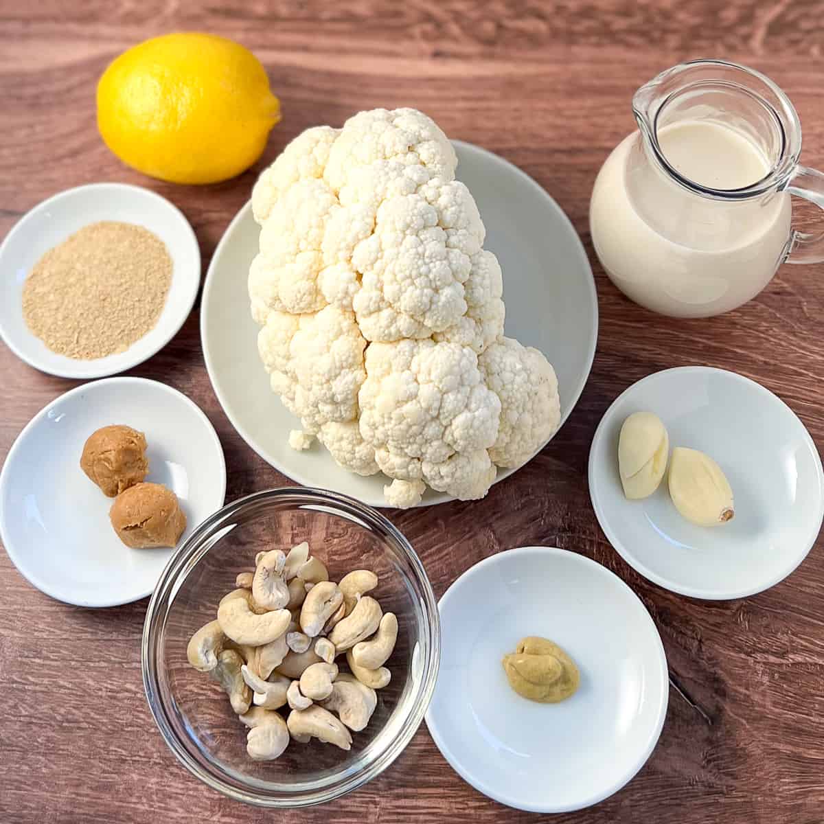 top view of ingredients for vegan alfredo sauce: cauliflower, cashews, miso paste, garlic, Dijon mustard, nutritional yeast, lemon and soymilk