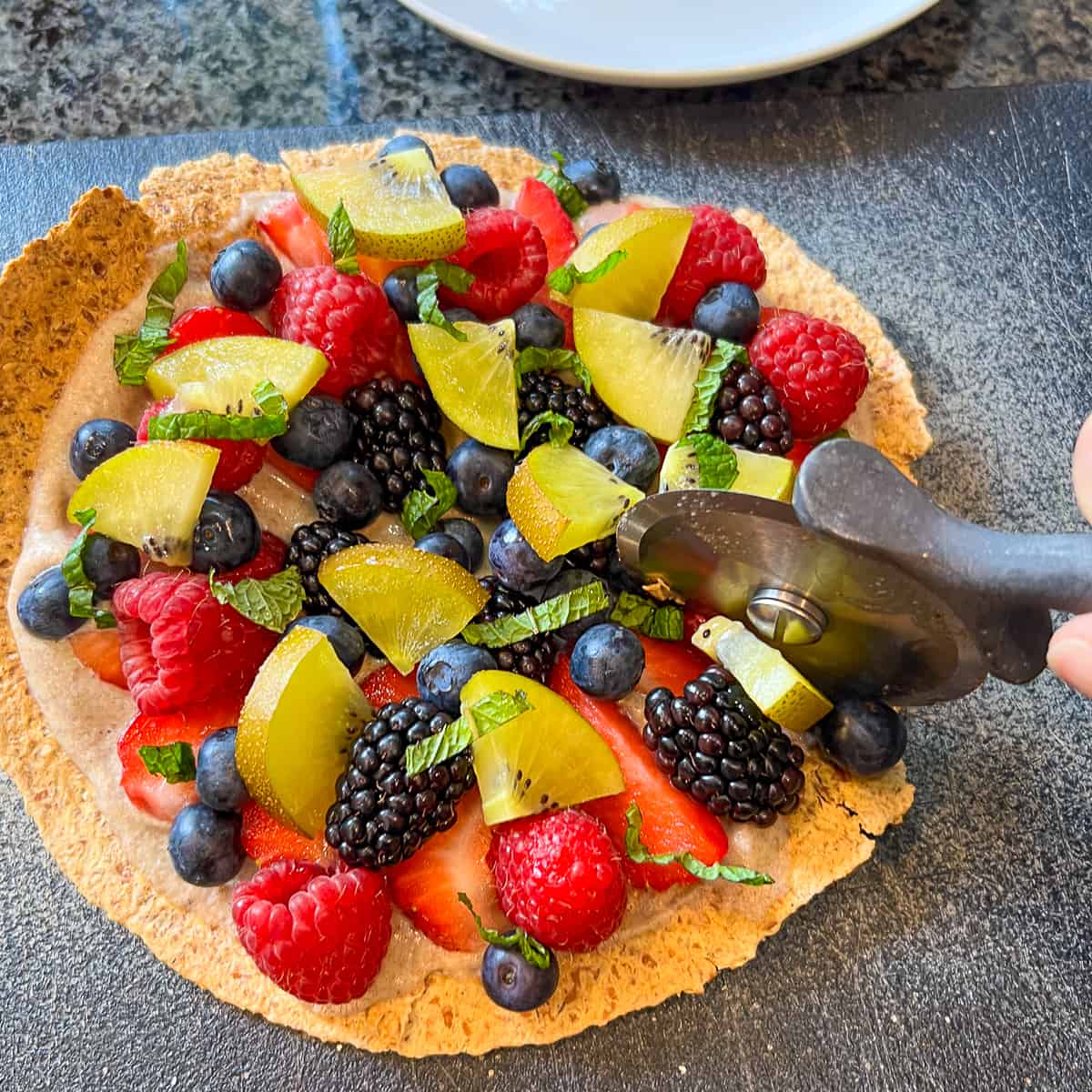 top view of fruit pizza being cut with a pizza cutter on a cutting board
