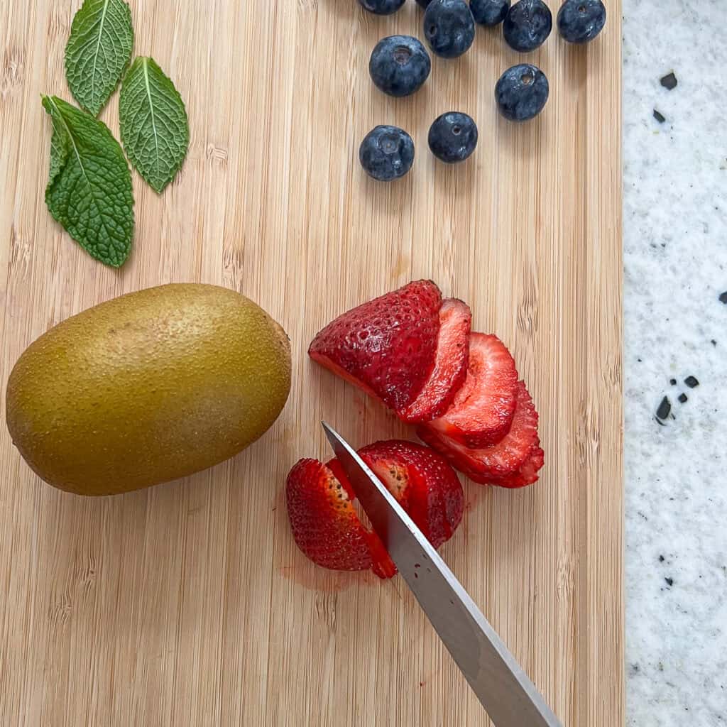 A knife slicing strawberries on a cutting board next to kiwi, mint and blueberries.