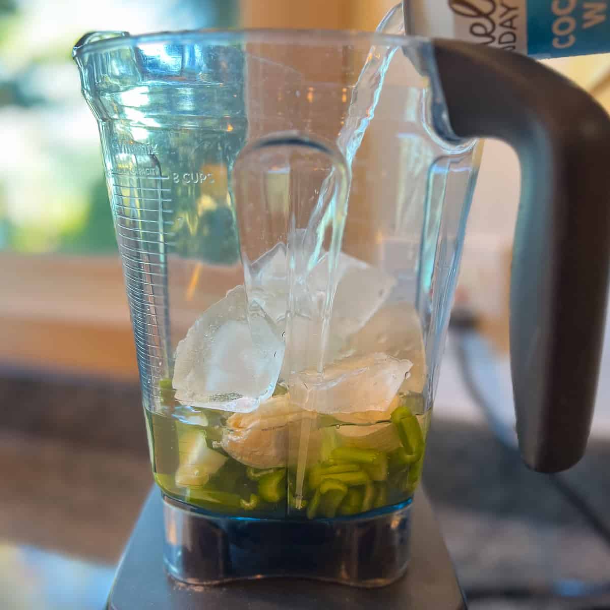 side view of celery, banana, cucumber, ice and coconut water being poured into a blender