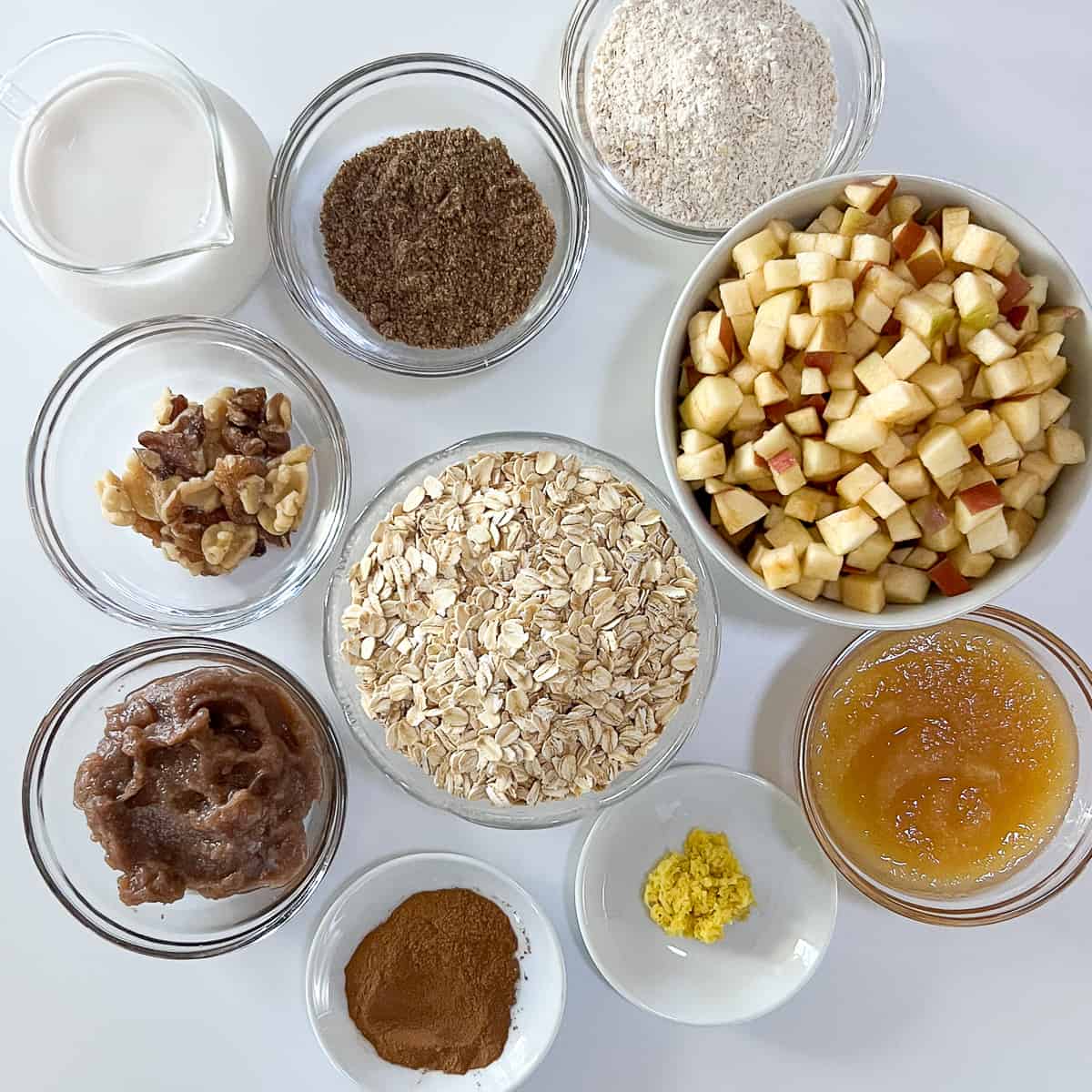 top view of ingredients for apple cinnamon muffins: diced apples, rolled oats, ground flaxseeds, apple sauce, walnuts, plant based milk, date syrup, grated ginger root, apple sauce and cinnamon
