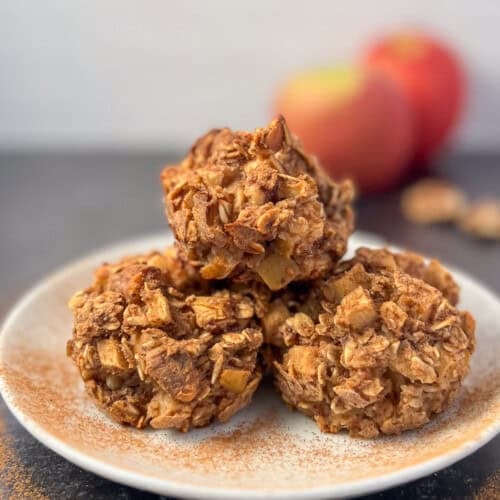 side view close up of three muffins stacked on a white plate with apples and walnuts blurred in the background