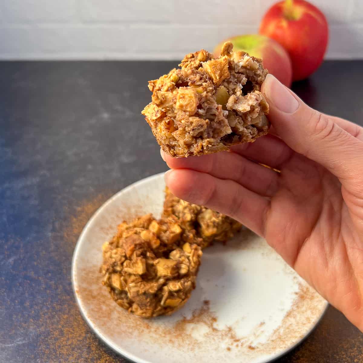 a woman's hand holding a partially eaten apple cinnamon oat muffin with other muffins and apples blurred in the background