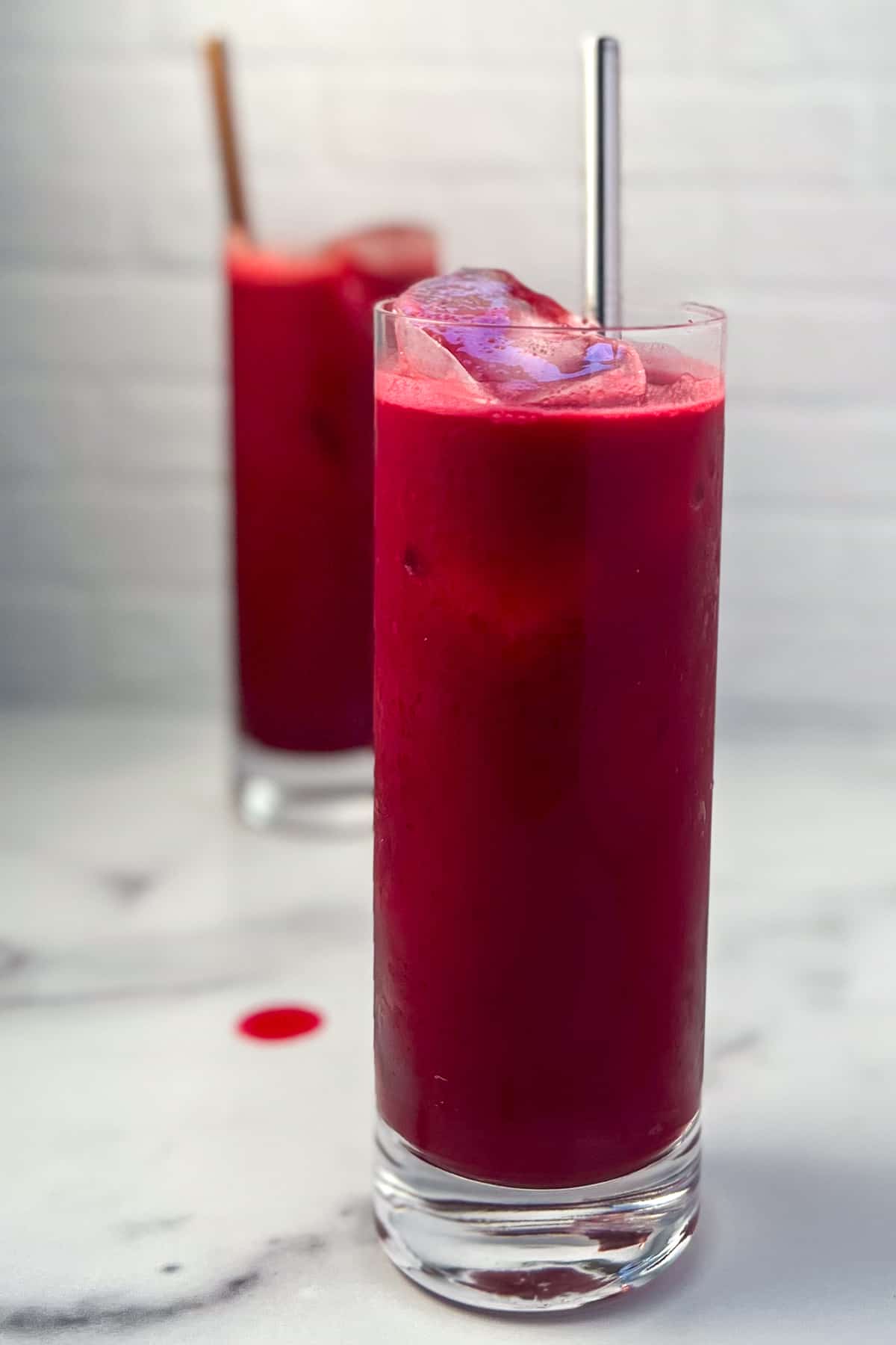 side view of tall glass of carrot beet apple juice with ice and metal straw sticking out; another glass of juice blurred in the background