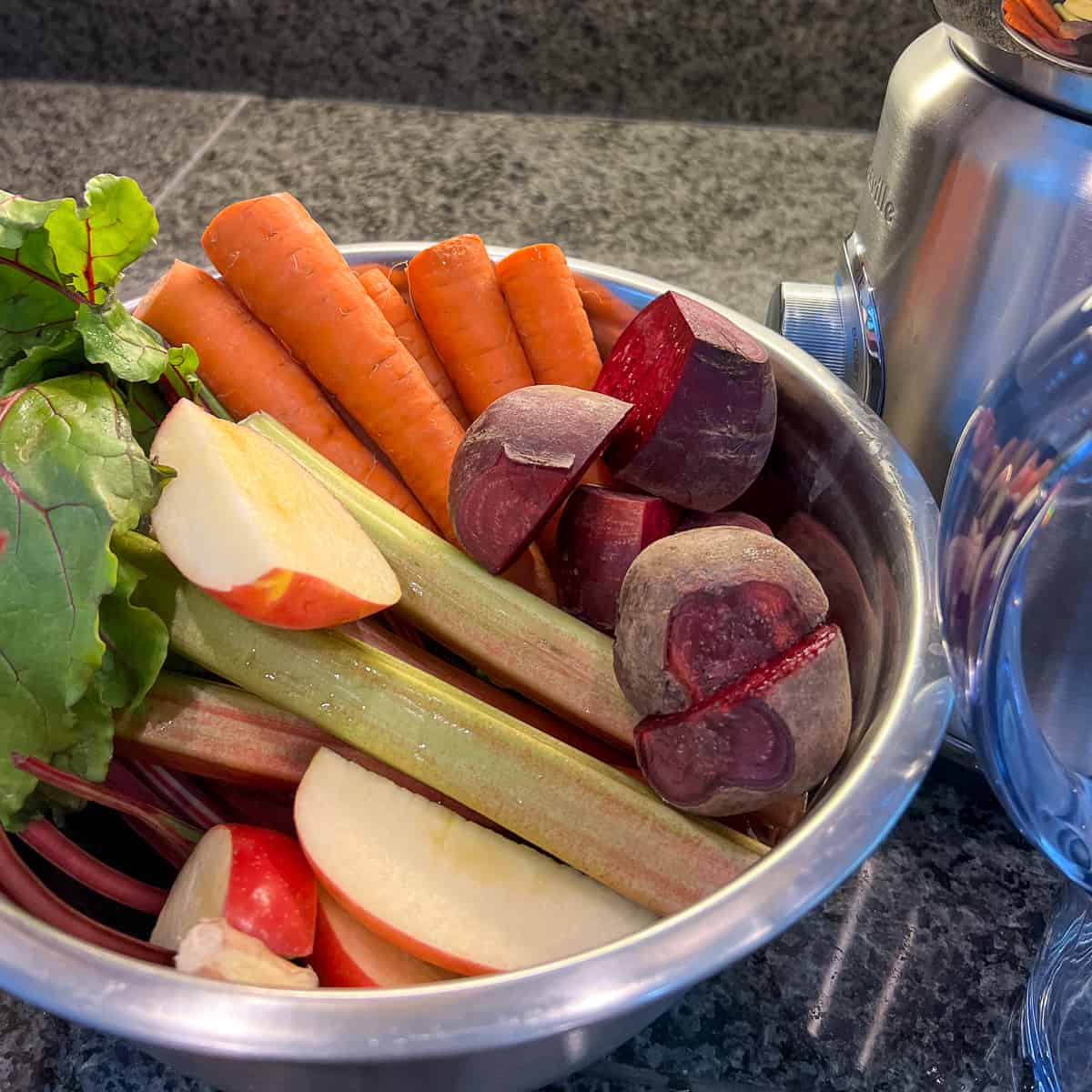 top side view of raw beets, carrots, rhubarb, apple and ginger in a large mixing bowl next to a stainless steel juicer