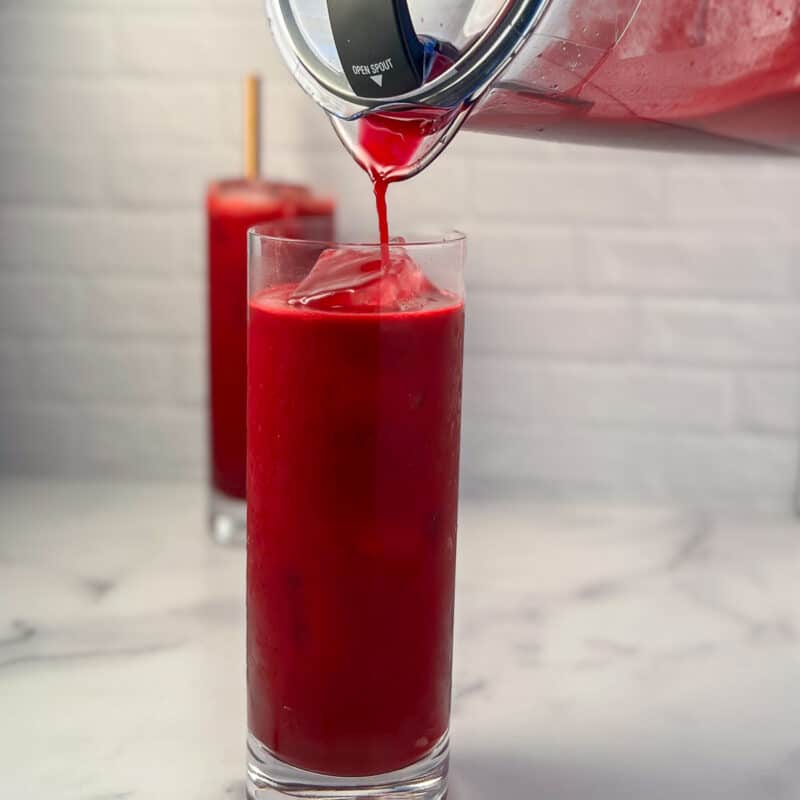 a pitcher pouring beet juice into a tall glass with ice; glass of beet juice blurred in the background
