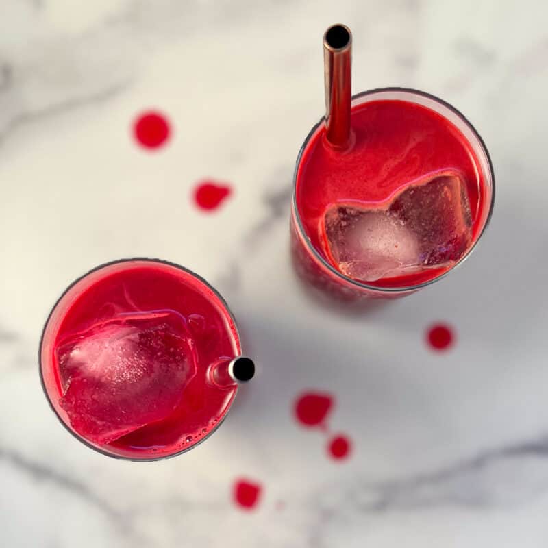 top view of two glasses of beet carrot apple juice in glass with ice and metal straws; splatters of juice on marble surface