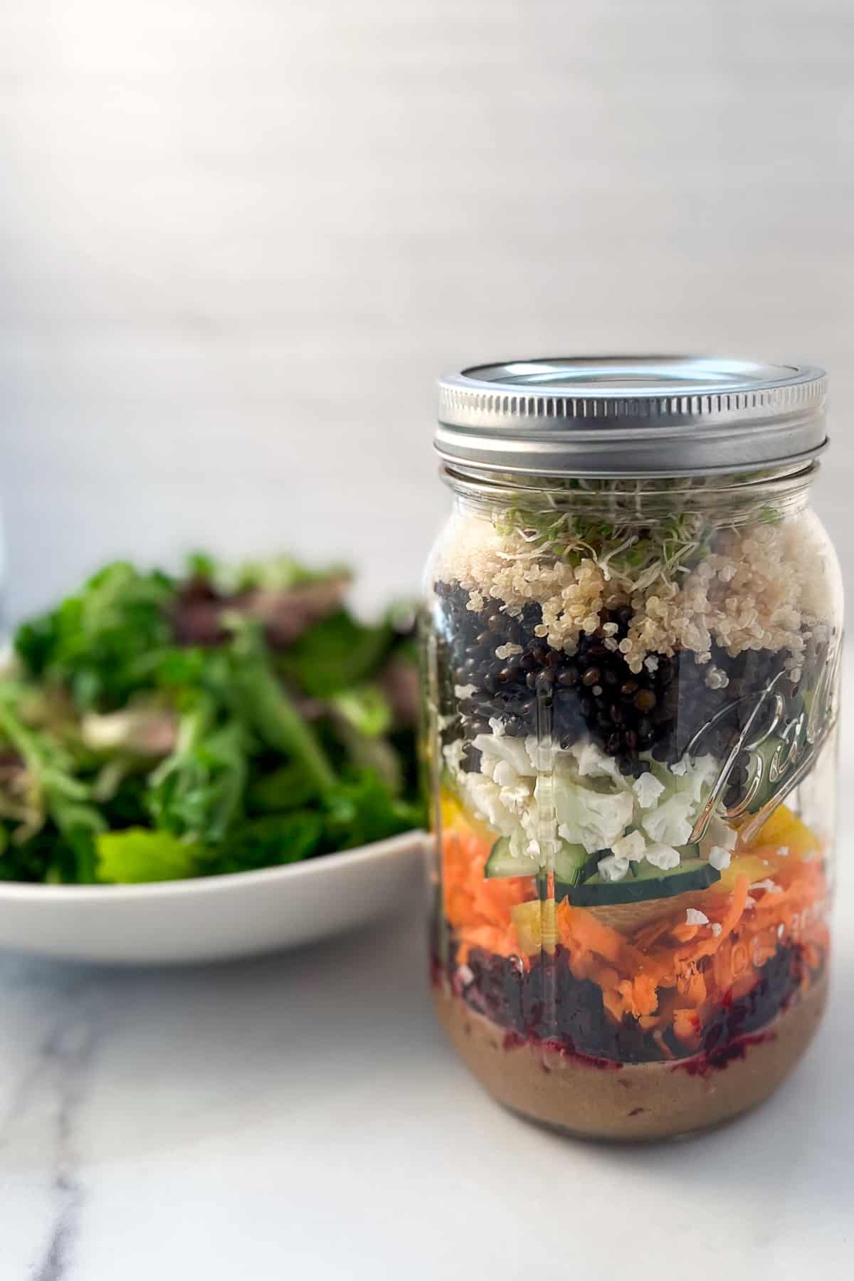 side view close up of superfood salad in a large mason jar and bowl of mixed salad greens blurred on the side