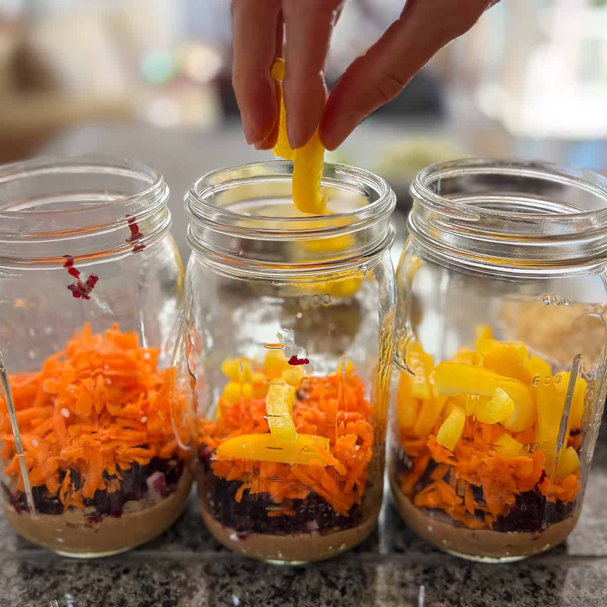 woman's hand adding chopped bell pepper to mason jars with other veggies