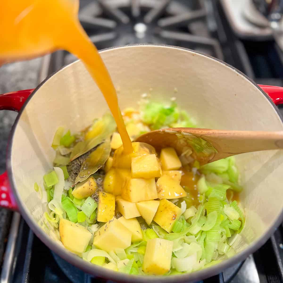 vegetable broth being added to the pot with leeks, potatoes, and spices on the stovetop