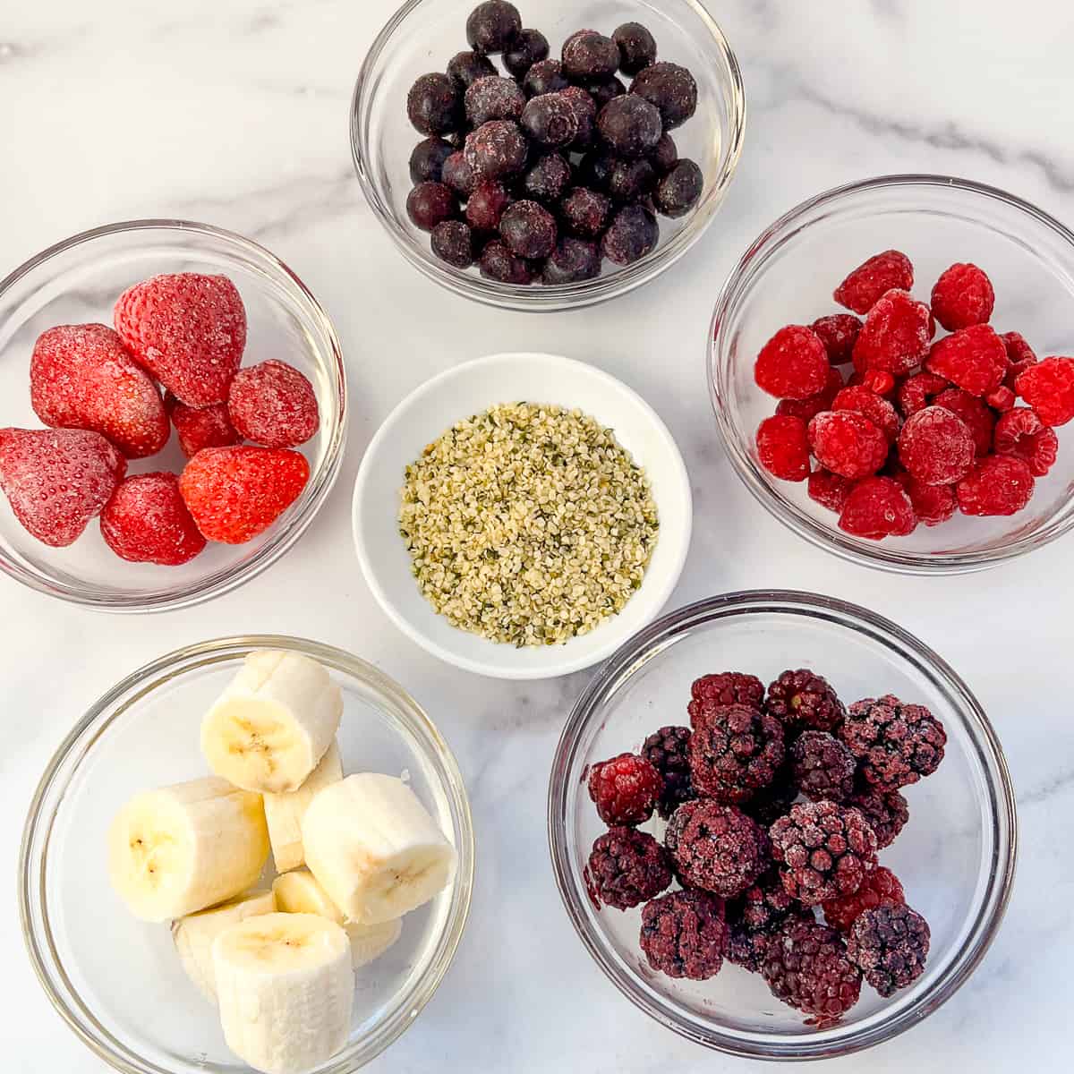 top view of key ingredients for mixed berry smoothie: frozen blueberries, frozen raspberries, frozen strawberries, frozen blackberries, banana and hemp hears