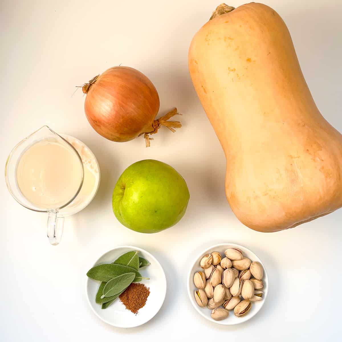top view of ingredients for butternut squash soup: butternut squash, onion, green apple, soymilk, ground nutmeg, fresh sage leaves and pistachios