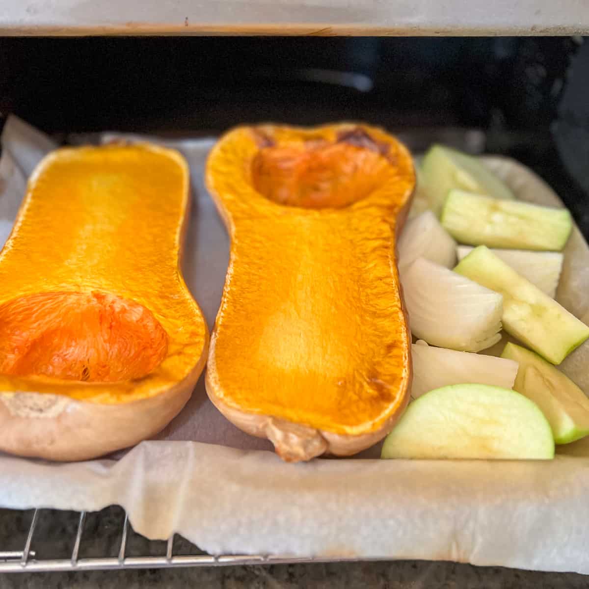 top side view of a baking sheet in the oven with butternut squash that's partially cooked on the left and uncooked raw onion and apple on the right