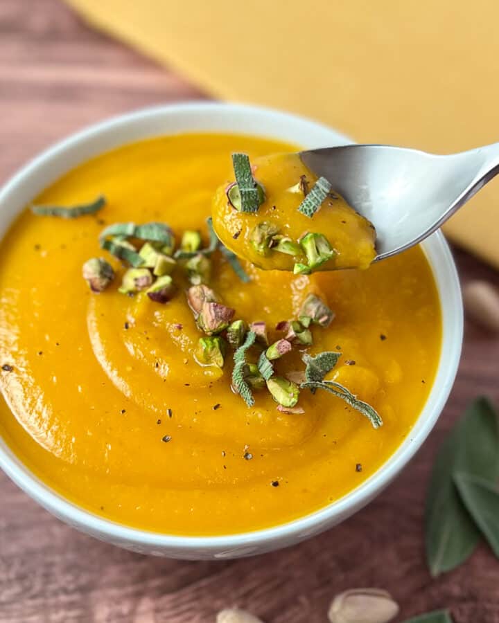 top side view of a bowl of vegan butternut squash soup topped with herbs and nuts and spoon dishing out some soup