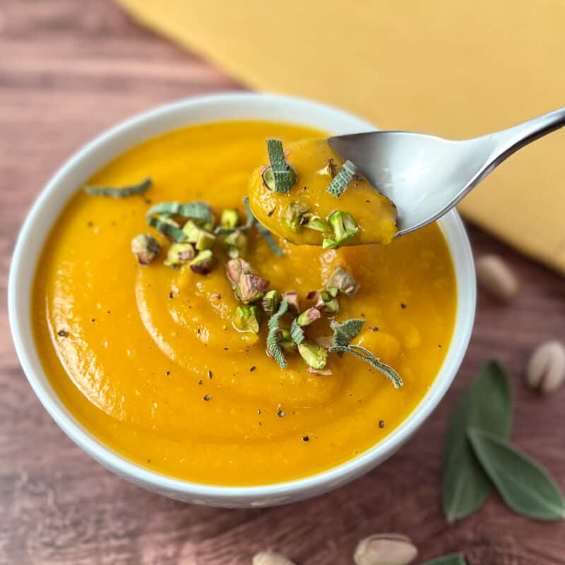 top side view of a bowl of vegan butternut squash soup topped with herbs and nuts and spoon dishing out some soup