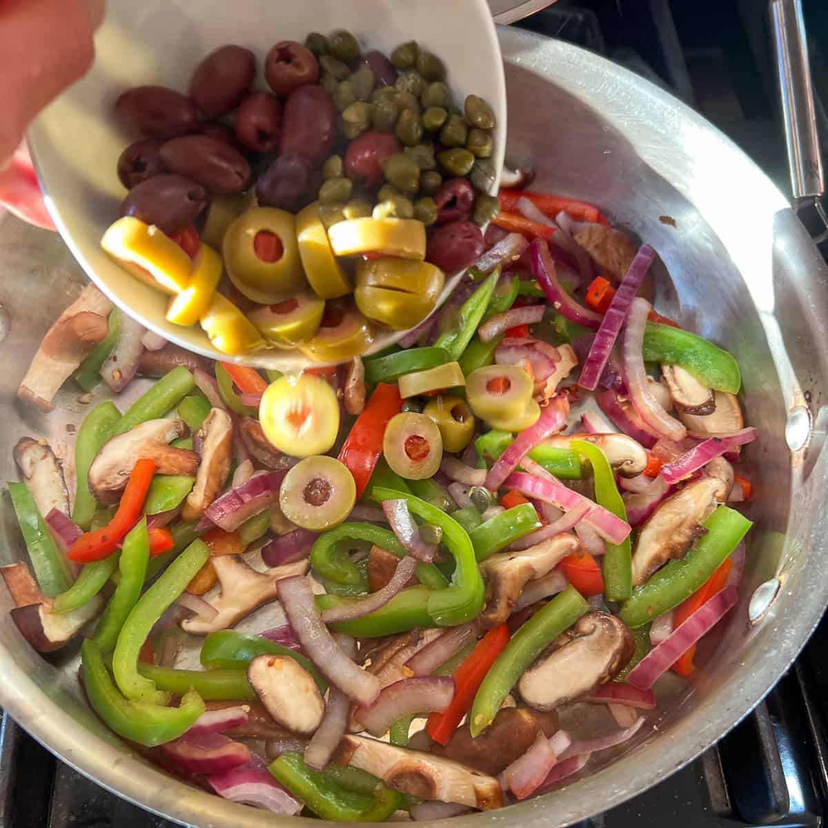olives and capers being added to a saute pan with veggies on the stovetop