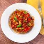 A plate of vegan puttanesca over spaghetti topped with fresh basil and a fork on the side.
