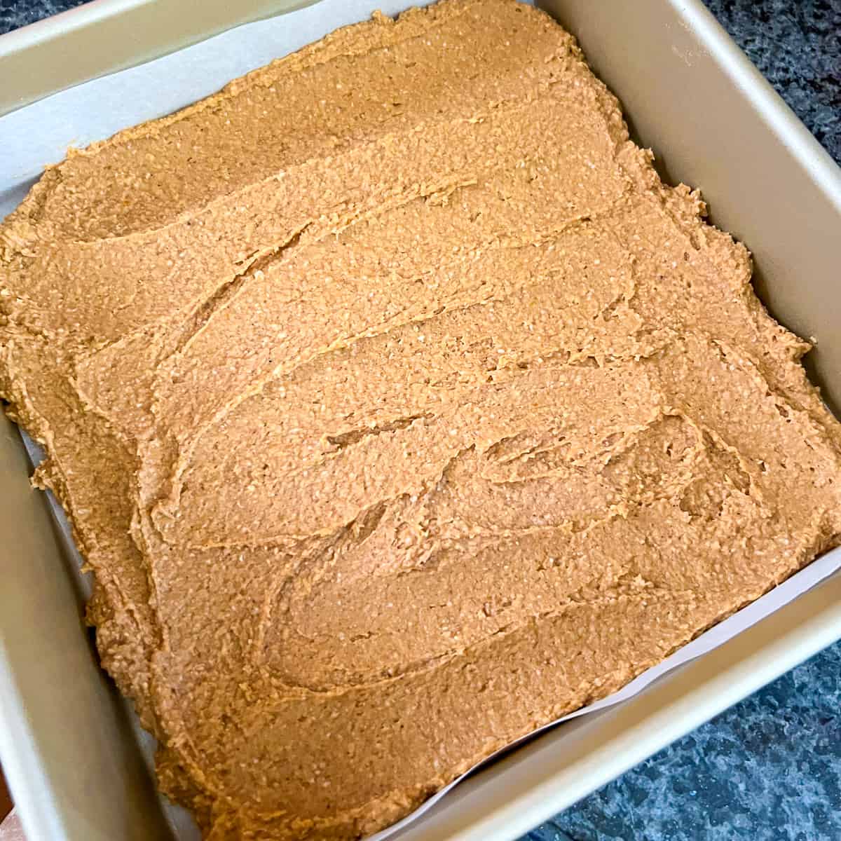 top view of a square pan with pumpkin brownie mixture spread evenly across the bottom of a parchment lined square baking pan