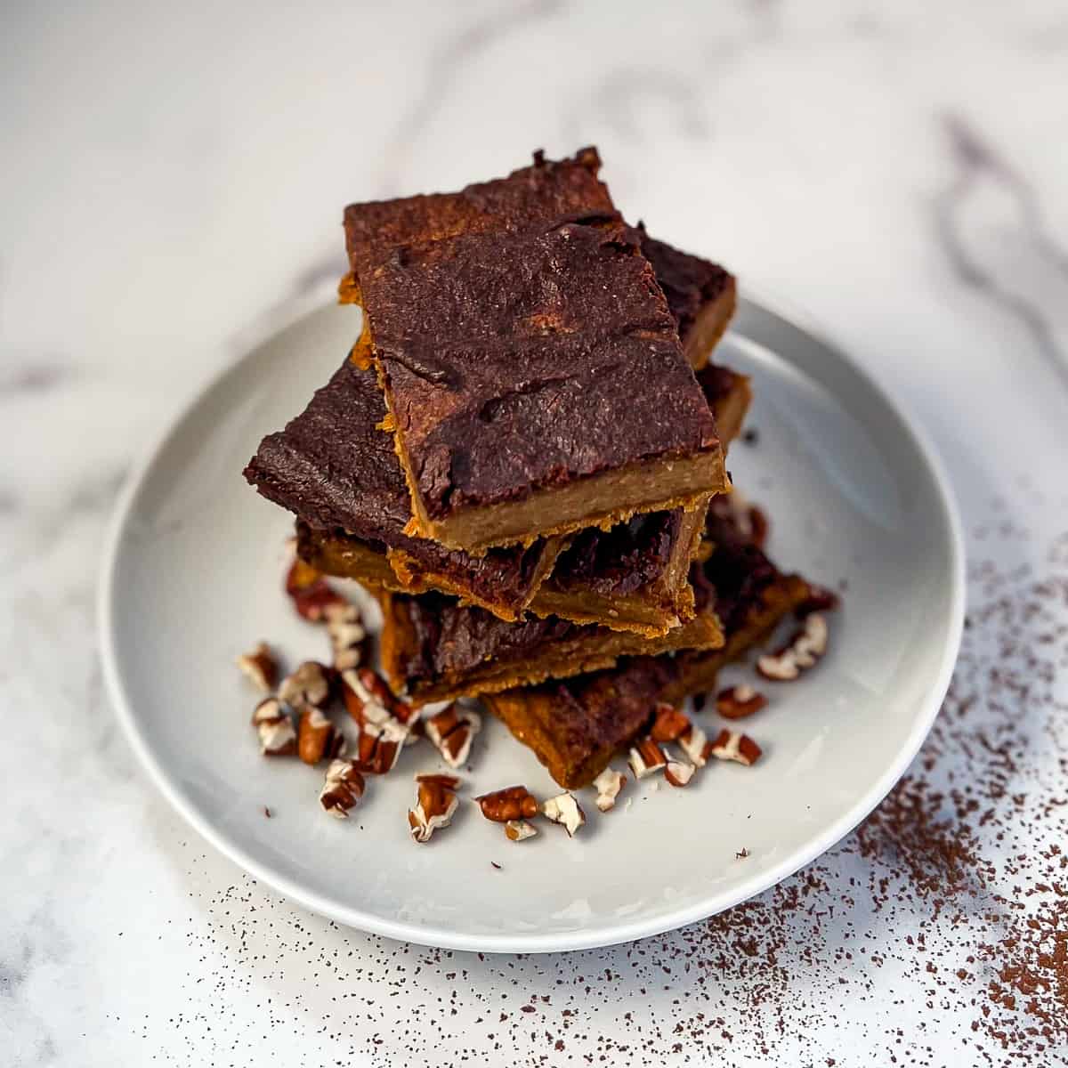 top side view close up of a stack of vegan pumpkin brownies on a white plate with chopped pecans and cocoa dust around it