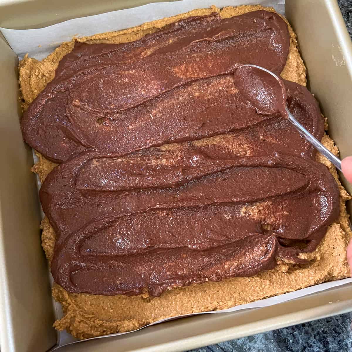 top view of chocolate tahini sauce being spread with a spoon across the top of a pan of pumpkin brownies