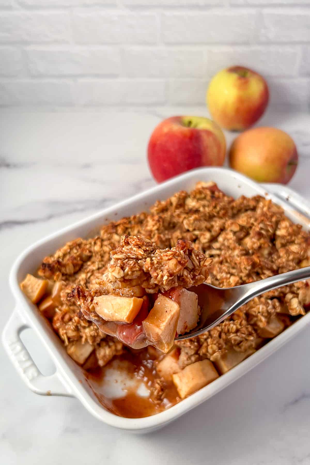 top side view of a vegan apple crisp in a casserole dish with spoon lifting some out; apples blurred in the background