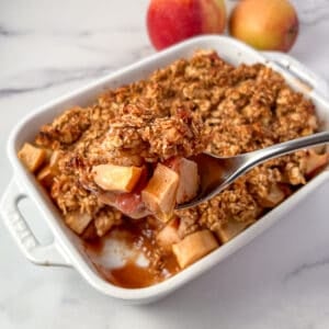 top side view close up of vegan apple crisp in a white casserole dish and spoon dishing some out; apples blurred in the background