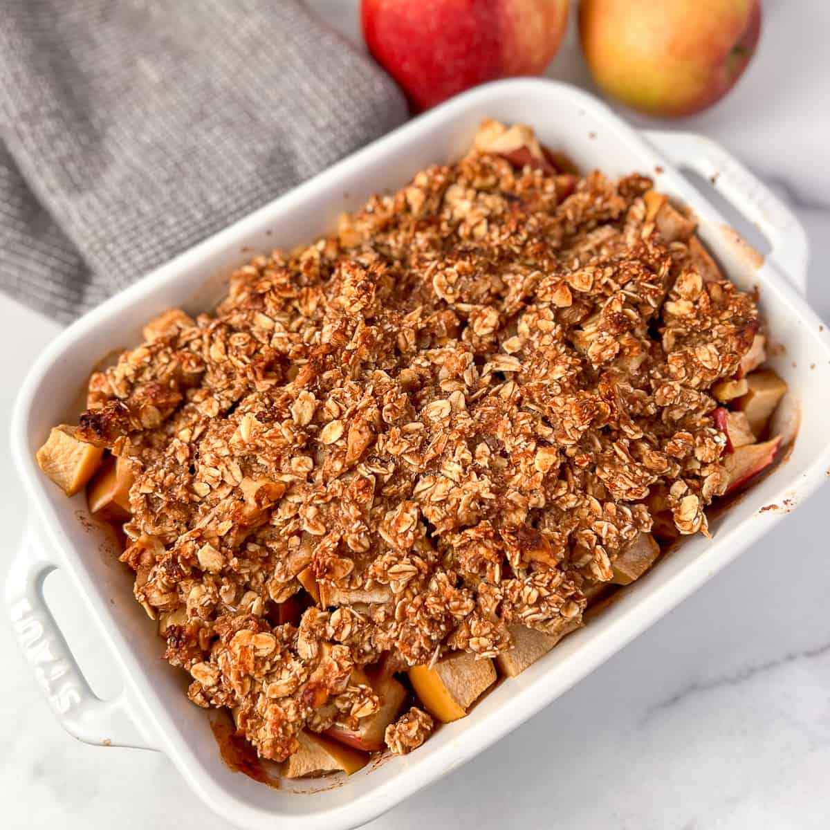 top view of vegan apple crisp in a white rectangular casserole dish; grey towel and apples blurred in the background