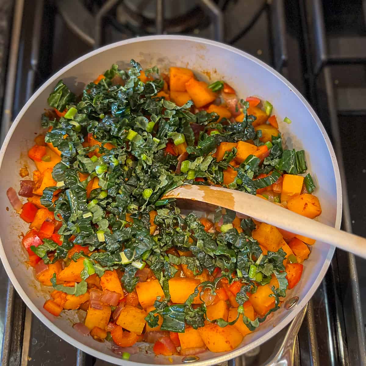 A wooden spoon stirring kale into the butternut squash hash in a pan on the stovetop.
