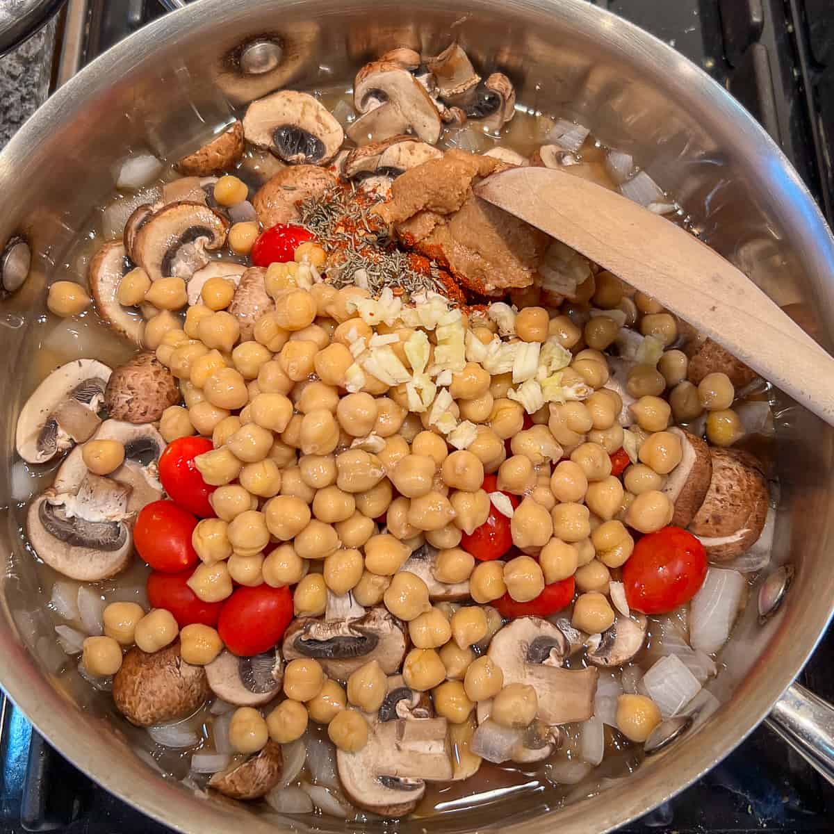 top view of chickpeas, tomatoes, garlic, miso paste and spices added to the saute pan with cooked onions and mushrooms