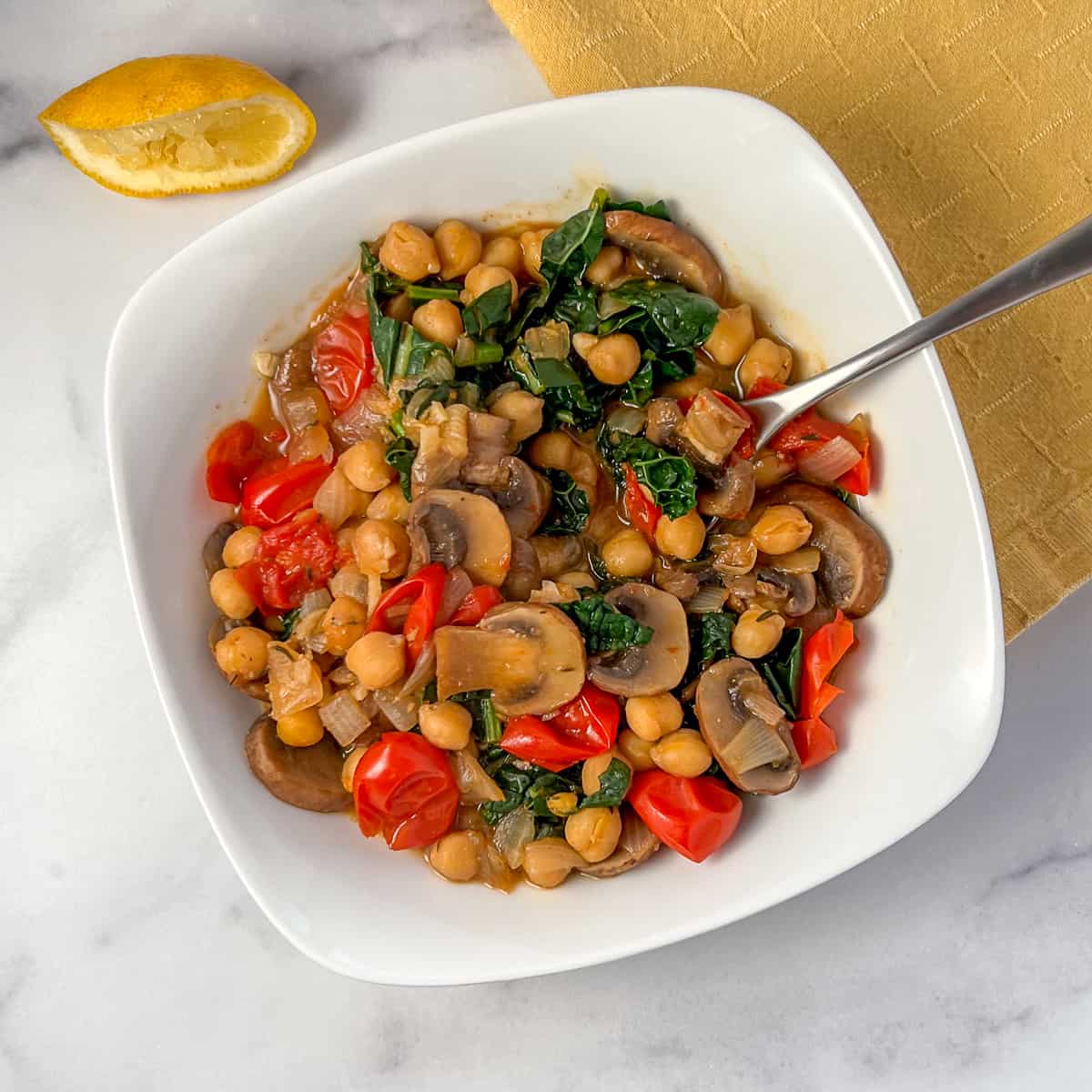 top view close up of vegan miso chickpea and mushroom stew in a white bowl with spoon; lemon and yellow napkin in background