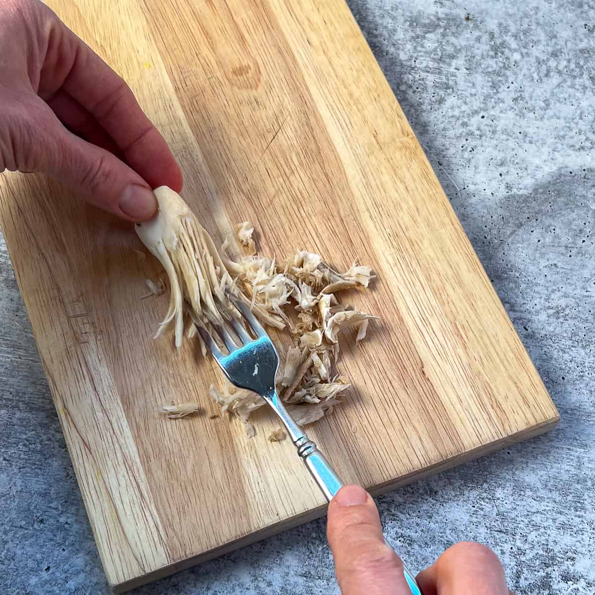 A woman's hands using a fork to shred the oyster mushrooms on a cutting board.